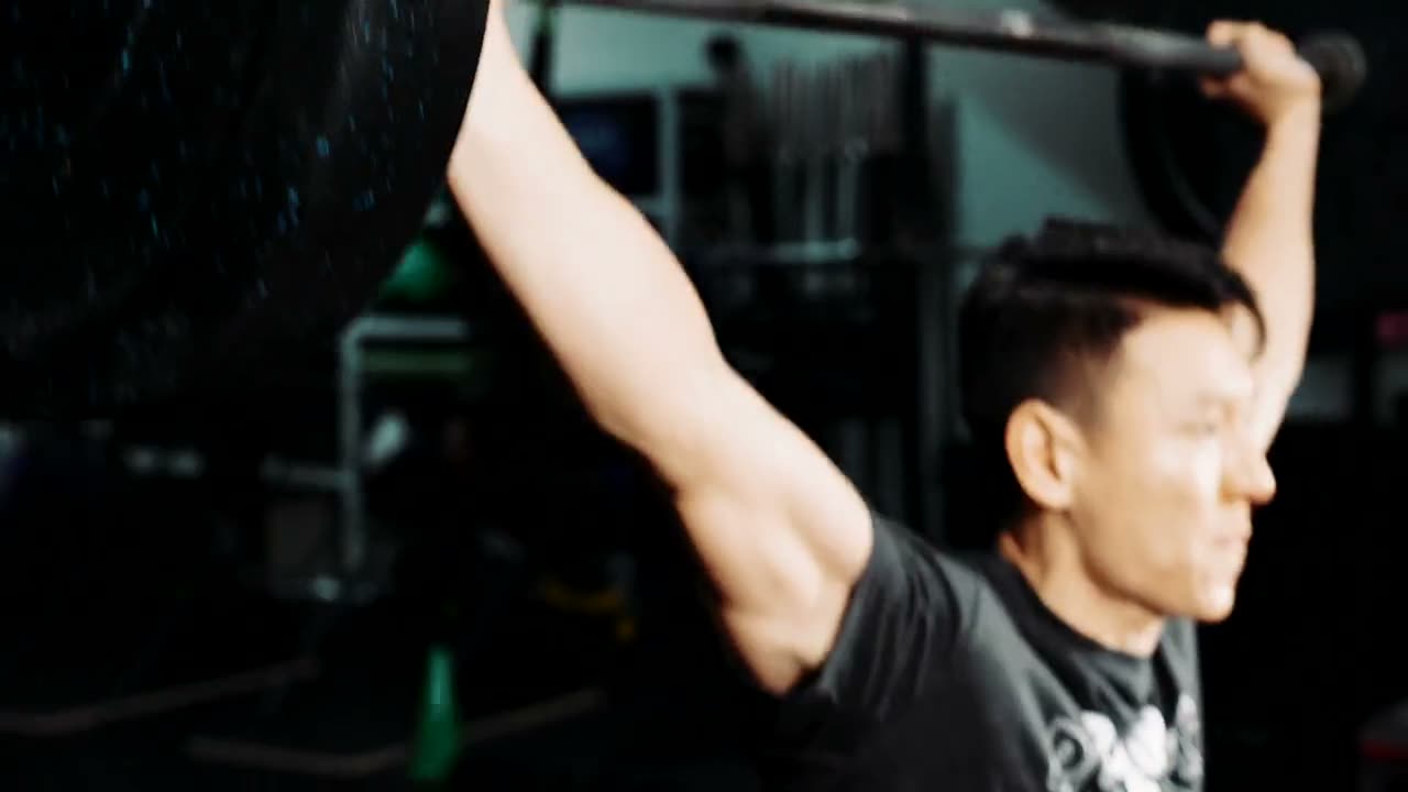 A lifter pressing a loaded barbell overhead in a dark gym, green plates on the bar.