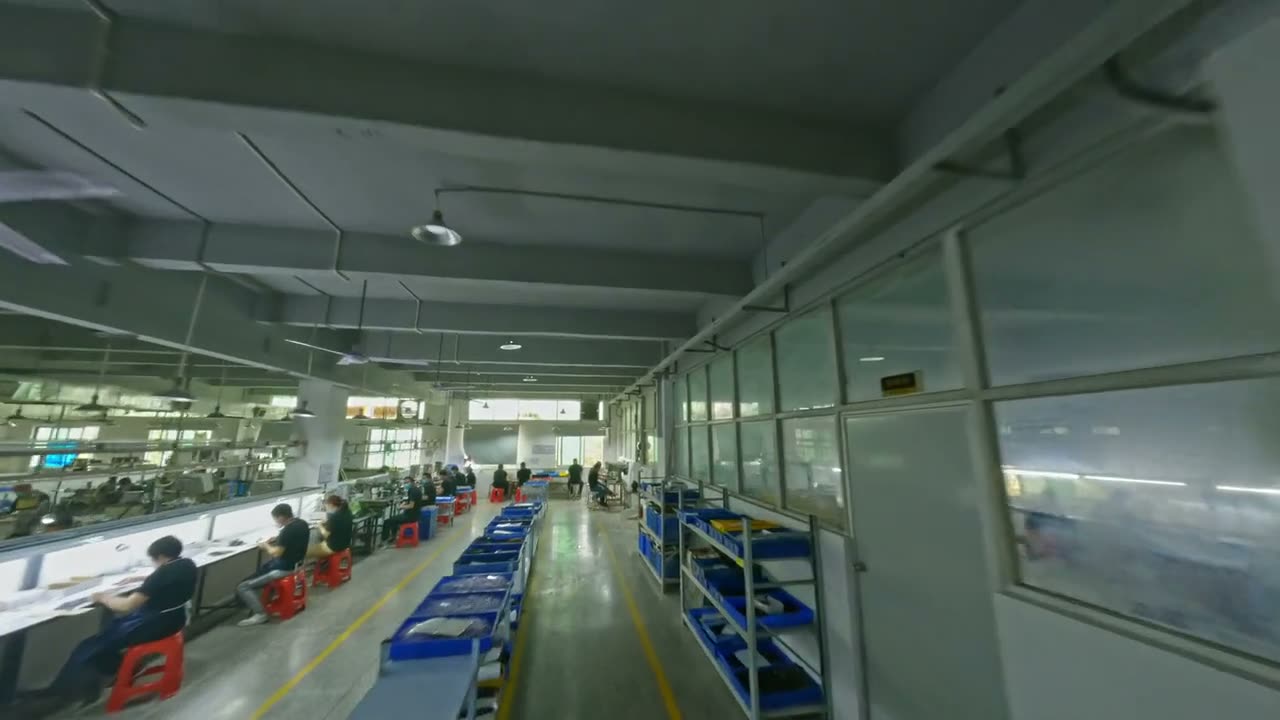 An FPV drone flies down the length of an assembly line with workers at benches on both sides.