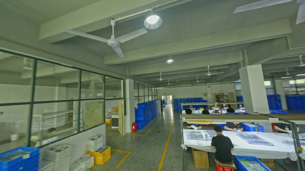 An FPV drone flies down an aisle of the LIBO factory, past benches where workers sit with rows of frames laid out in front of them.