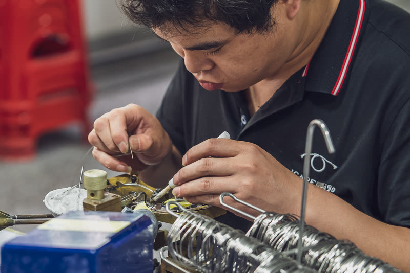 A technician solders a wire frame at the bench, a rack of formed fronts alongside.