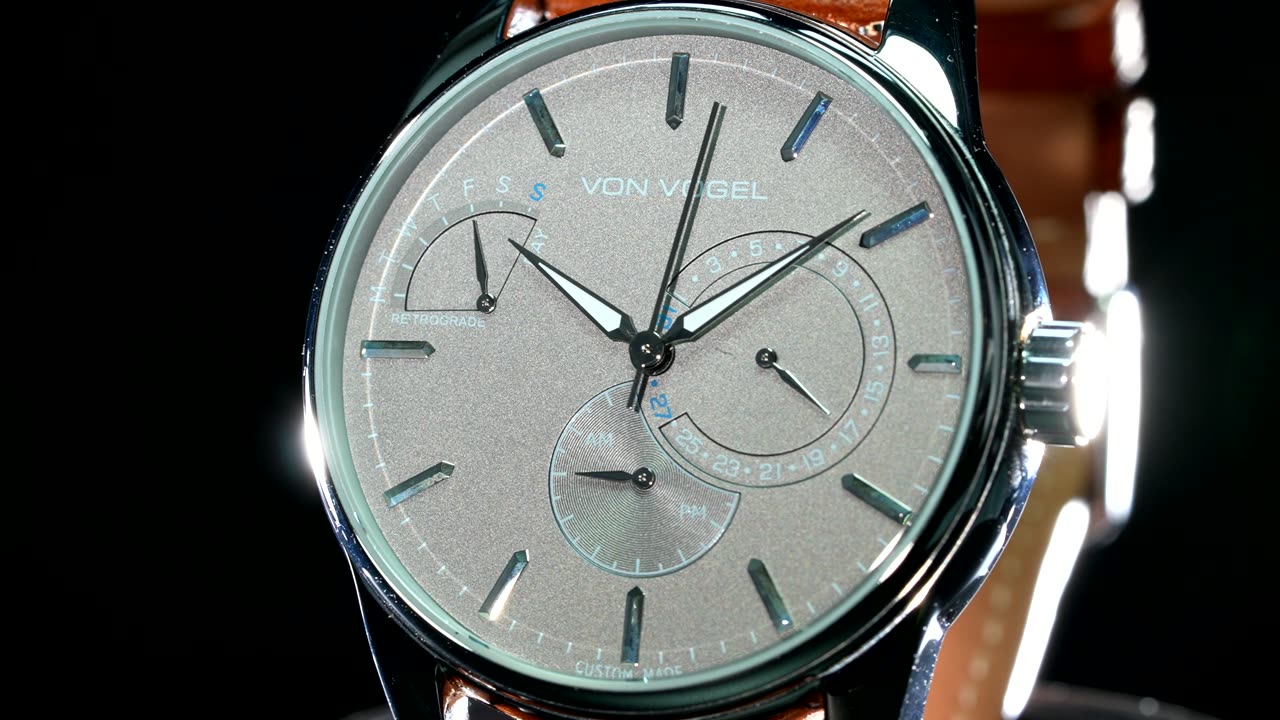 A silver Von Vogel dial with a retrograde day arc, lit against black.