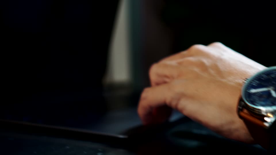 The watch at a desk on a brown leather strap, at the cuff of a hand resting on a laptop.