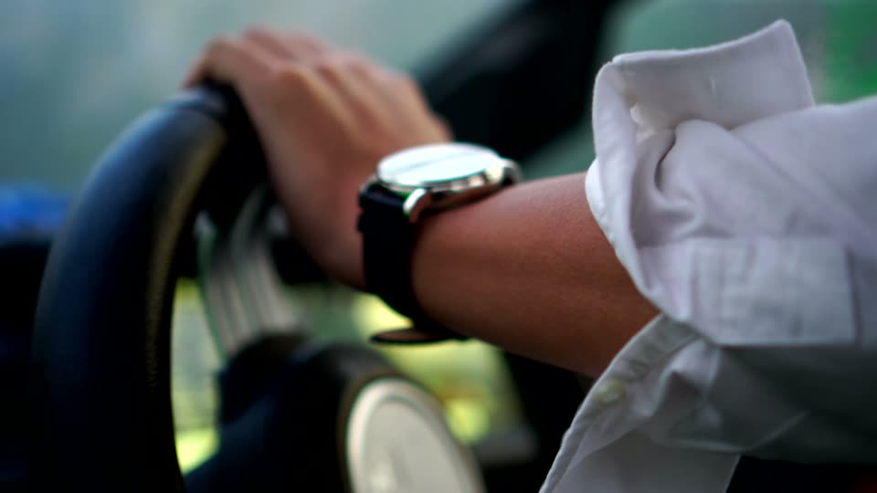 The watch on a black strap at a white shirt cuff, a hand on a steering wheel.